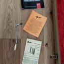 Na dřevěné podlaze leží oranžový manuál s nápisy „OWNER'S MANUAL for Stratocaster and Telecaster“ a „Crafted in the American Tradition“ spolu s logem…