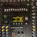 Detailní pohled na míchací pult stříbrné barvy s nápisem Dynacord CMS 2200 COMPACT MIXING SYSTEM. Uprostřed je umístěn digitální displej s texty BRIG…