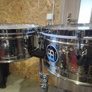 Dvě stříbrné bicí bubny s povrchem v optice kovaného kovu. Na pravém bubnu je nalepená modro-bílá kulatá nálepka s nápisem „soundhouse“ a stylizovaný…