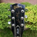 Fotografie zachycuje hlavu černé elektrické kytary s chromovanými ladičkami. Na povrchu je zlatým písmem nápis Gibson, Les Paul MODEL a Studio. Lak n…
