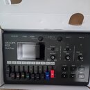 Černý digitální multitrackový záznamník s nápisy ZOOM, R12 MultiTrak. Zařízení má displej, několik ovládacích prvků a osm faderů s barevnými značkami…