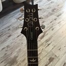 Fotografie zachycuje hmatník a hlavu elektrické kytary v tmavě hnědé barvě. Na hlavě je bílý nápis "Paul Reed Smith" v rukopisném stylu a na spodní h…