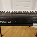 Černý digitální klavír umístěný na černém stojanu. Na přední straně je bílý nápis JUNO-D. V pravé části jsou vidět konektory s popisky MIDI OUT, MIDI…