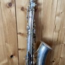 Stříbrný saxofon s kovovým povrchem, který vykazuje známky opotřebení a oxidace. Nástroj je vybaven světle hnědým mouthpiecem. Na zvonu jsou viditeln…