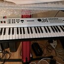 Bílá klávesová syntezátor s černými klávesami umístěná na červeném stojanu. Na levé straně je nápis YAMAHA a v pravém horním rohu označení MX61. Horn…
