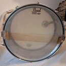 Fotografie znázorňuje basový buben s průhledným koženem. Na horní části kožeje je černý nápis Pearl a pod ním text AMBASSADOR DRUM HEAD BY REMO U.S.A…