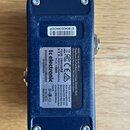 Zadní strana modrého elektronického zařízení v plastovém nebo kovovém krytu s čtyřmi viditelnými šrouby v rozích. Na zařízení jsou dvě nálepky; horní…