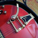 Detail červené elektrické kytary s instalovaným zlatým tremolo mechanismem. Na kovovém krytu mechanismu je viditelné logo "bigsby" a nápis "LICENSED"…