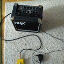 Černý kytarový kombo zesilovač s nápisy MIGHTY 8 a NUX. K zesilovači je připojený žlutýstupač s logem NUX a napájecí adaptér. Vše je umístěno na bílé…