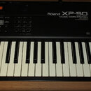 Černý klávesový syntezátor s bílými a černými klávesami. Na horním panelu je nápis Roland XP-50 MUSIC WORKSTATION 16 VOICE 4x EXPANSION a logo MIDI.…
