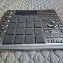 Stříbrný MPC sampler s mřížkou šestiдесяti čtyř šedých padů a několika ovládacími knoflíky vlevo. V pravém horním rohu je červené logo AKAI a pod dis…