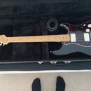 Černá elektrická kytara s červeně vzorovaným pickguardem v černém tvrdém kufru. Na hlavě hmatníku je viditelné logo Fender. Nástroj má světle hnědý j…