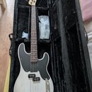 Elektrická basová kytara s bílým trupem a černým pickguardem v černém měkkém pouzdře. Na hlavě hmatníku je viditelné logo Fender. Nástroj má hnědý hm…