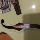 Detail korpusu světle zelené kytary s f-otvorem. Uvnitř otvoru je vidět štítek s nápisy Model No. G6118T-LTV a Serial No. JT08030841. Na povrchu laku…