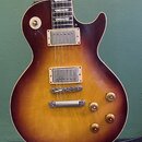 Elektrická kytara s trupem v barvě sunburst (přechod od žluté k tmavě hnědé). Nástroj má dva humbuckery s kovovými kryty, čtyři zlaté oválné knofíky…