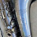 Detail stříbrného saxofonu s viditelným opotřebením povrchu a drobnými škrábanci. Na trubici jsou vyryty nápisy „H. Selmer“, „4. PLACE DANCOURT PARIS…