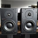 Dva černé regálové reproduktory umístěné na černých stojanech. Na přední straně každého reproduktoru je nápis „dynaudio acoustics“ na červeném pozadí…