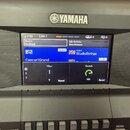 Detailní pohled na ovládací panel klávesového nástroje značky YAMAHA. Uprostřed je zapnutý displej, na kterém jsou vidět nápisy Regist NewBank, Mic S…