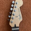 Fotografie zachycuje hlavu elektrokitary z přírodního světlého dřeva s chromovanými ladičkami. Na hlavě je černé logo Fender, nápis STRATOCASTER a te…