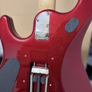 Zadní strana červené elektrické kytary s kovým krytem krkovým spojem. Na krytu je vyražen nápis ERNIE BALL MUSIC MAN a sériové číslo 600073. V dolní…