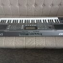 Šedý digitální klavír s bílými a černými klávesami. Na přední hraně je vlevo nápis Roland a vpravo označení EXR-5s. Na horní části těla je vpravo log…