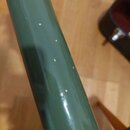 Detailní záběr na tmavě zelený, lakovaný povrch trubičky hudebního nástroje. Na povrchu je vidět několik drobných odlakování a světlých škrábanců. Žá…