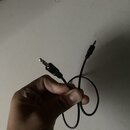 Černý audio kabel s jack konektory na obou koncích. Kabel je v zakrouceném tvaru a drží ho ruka. Na konektorech nejsou viditelné žádné značky ani náp…