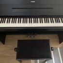 Černý digitální piano s klaviaturou a přívodním kabelem. Na horní části korpusu je bílé logo YAMAHA. Součástí je černá pianová stolička s polstrování…