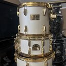 Tři bílé bubny s pozlaceným hardwarem naskládané na sebe. Na každém je viditelný štítek s nápisem Pearl Masters Custom. Povrch nástrojů i kovové část…