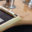 Detailní záběr na horní část hmatníku a hlavu kytary. Vidět jsou bílé pražce osazené na tmavém dřevě s perleťovou vložkou. Na hraně hmatníku je světl…