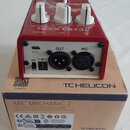 Červený efektový pedál TC HELICON MIC MECHANIC 2 umístěný na své originální kartonové krabici. Na horní straně jsou tři bílé oválové ovladače a nápis…
