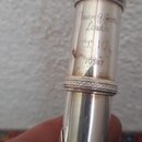 Detail horní části stříbrného dechového nástroje. Na povrchu je vyryto „Trevor G. James London“, pod tím značka „T.J. 10%“ a sériové číslo „70387“. P…