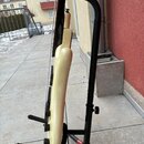 Bílá elektrická basa v černém stojanu. Nástroj má hladký lakovaný povrch a černé ovládací knoflíky. Na fotce nejsou žádné viditelné značky ani nápisy.