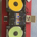 Sada DJ vybavení složená ze dvou černých gramofonů a centrálního mixeru, umístěná na červeném povrchu. Horní gramofon má žlutý vinyl, dolní světle ze…