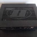 Černý elektronický přístroj s kovovým šasi a ventilační mřížkou na horní straně. V mřížce je vyřezáno logo znázorňující dvě svislé čáry v kruhu. Zaří…