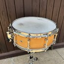 Dřevěný snare drum s přirozeným odstínem dřeva a chromovaným kováním. Na blaně je viditelné logo REMO. Povrch blány vykazuje známky používání ve form…