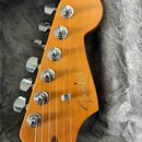 Detail hlavy elektrické kytary z přírodního dřeva s lakovaným povrchem. Na hlavě je umístěno logo Fender a nápis STRATOCASTER. Jsou vidět šest chromo…