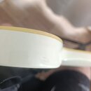 Detailní pohled na hranu bílého trupu hudebního nástroje s žlutým lemováním. Povrch je hladký a glossy, v dolní části jsou vidět drobné škrábance. Žá…