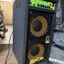 Černý basový kombozesilovač s potahem z textilie a plastovými rohy. Horní ovládací panel je zelený s nápisy Markbass a Little Mark Ninja. Na spodní č…
