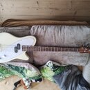 Elektrická basová kytara v krémové barvě s bílým perleťovým pickguardem. Na hlavě hnedbového krku je viditelné logo "Fender". Nástroj má čtyři struny…