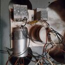 Fotografie zachycuje vnitřní zapojení elektroniky s několika komponenty na bílém podkladu. Jsou zde vidět dva kovové transformátory, jeden velký hlin…