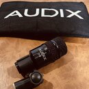 Černý dynamický mikrofon s mřížkou, na kterém je bílý nápis AUDIX D4. Mikrofon je upevněn v černém držáku s plastovým šroubem pro utahování. V pozadí…