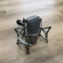 Stříbrný kondenzátorový mikrofon s kovovou síťkou, umístěný v odpružíného držáku stejné barvy. Na spodní části těla mikrofonu je červené logo s nápis…