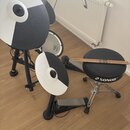Elektronická bicí souprava s černými stojany a bílo-černými pady. Na padu činelů i na basovém pedálu je viditelný nápis Roland. Na černém sedadle je…