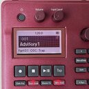 Detailní pohled na ovládací panel červeného elektronického nástroje. Uprostřed je displej s nápisy 128.0, 001, Advisory 1 a Part:01 OSC: Trap. Nad di…