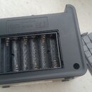 Zadní strana černého elektronického zařízení s otevřeným přihrádkou pro baterie, ve které jsou vloženy čtyři cylindrické články. V horní části je vyr…