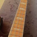 Fotografie zachycuje hmatník a hlavu basové kytary s dřevěným povrchem a světle barevnými obdélníkovými vložkami místo pražcových značek. Na hlavě ná…