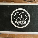 Černý transportní kufr s hliníkovým rámem a kovovými zapínacími prvky. Uprostřed víka je bílé kulaté logo s nápisem AKG a třemi překrytými ovály. Pov…