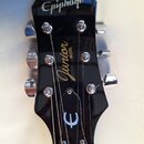 Fotografie zachycuje hlavu elektrické kytary v černé barvě s chromovanými ladičkami. Na horní části je bílý nápis Epiphone, uprostřed zlatý nápis Jun…