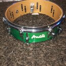 Zelený snare drum s kovovými obručemi a mechanismem na napnutí blikačů. Na korpusu je bílý nápis Amati. Nástroj vykazuje známky používání a drobné od…